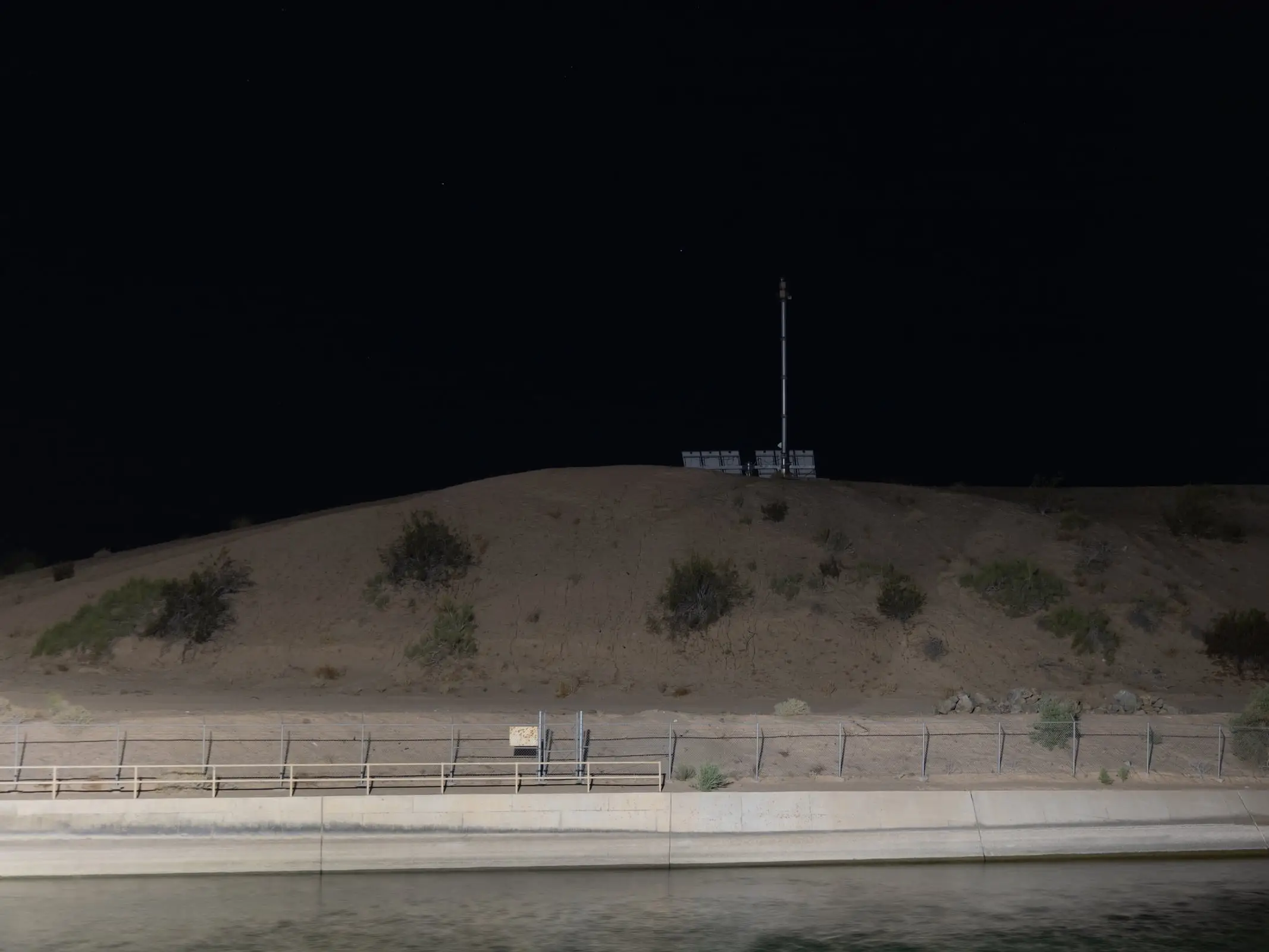 A solar-powered surveillance tower on a ridge above a border canal at night