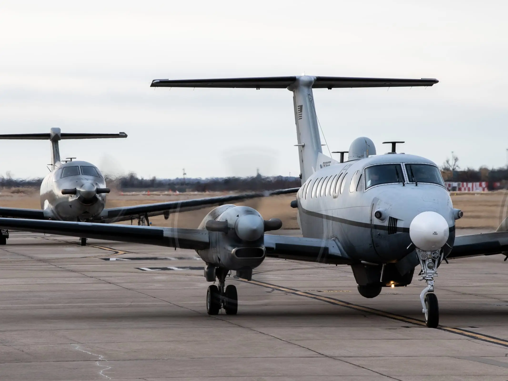 Two King Air airframes in low-visibility gray on a taxiway, the nearer one carrying a dorsal fairing and a belly sensor