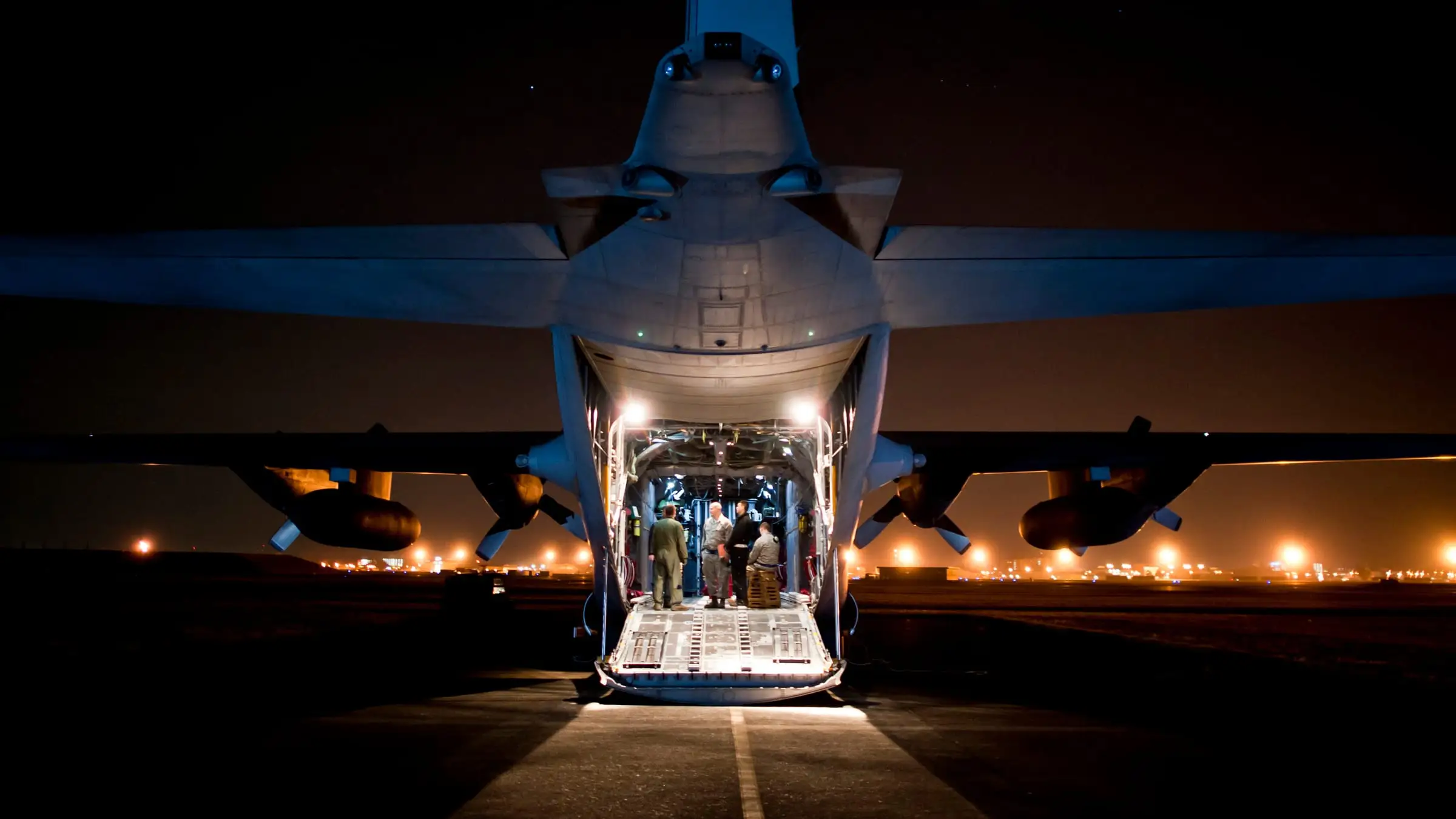 A transport aircraft on a floodlit apron at night, ramp down and cargo bay lit from within