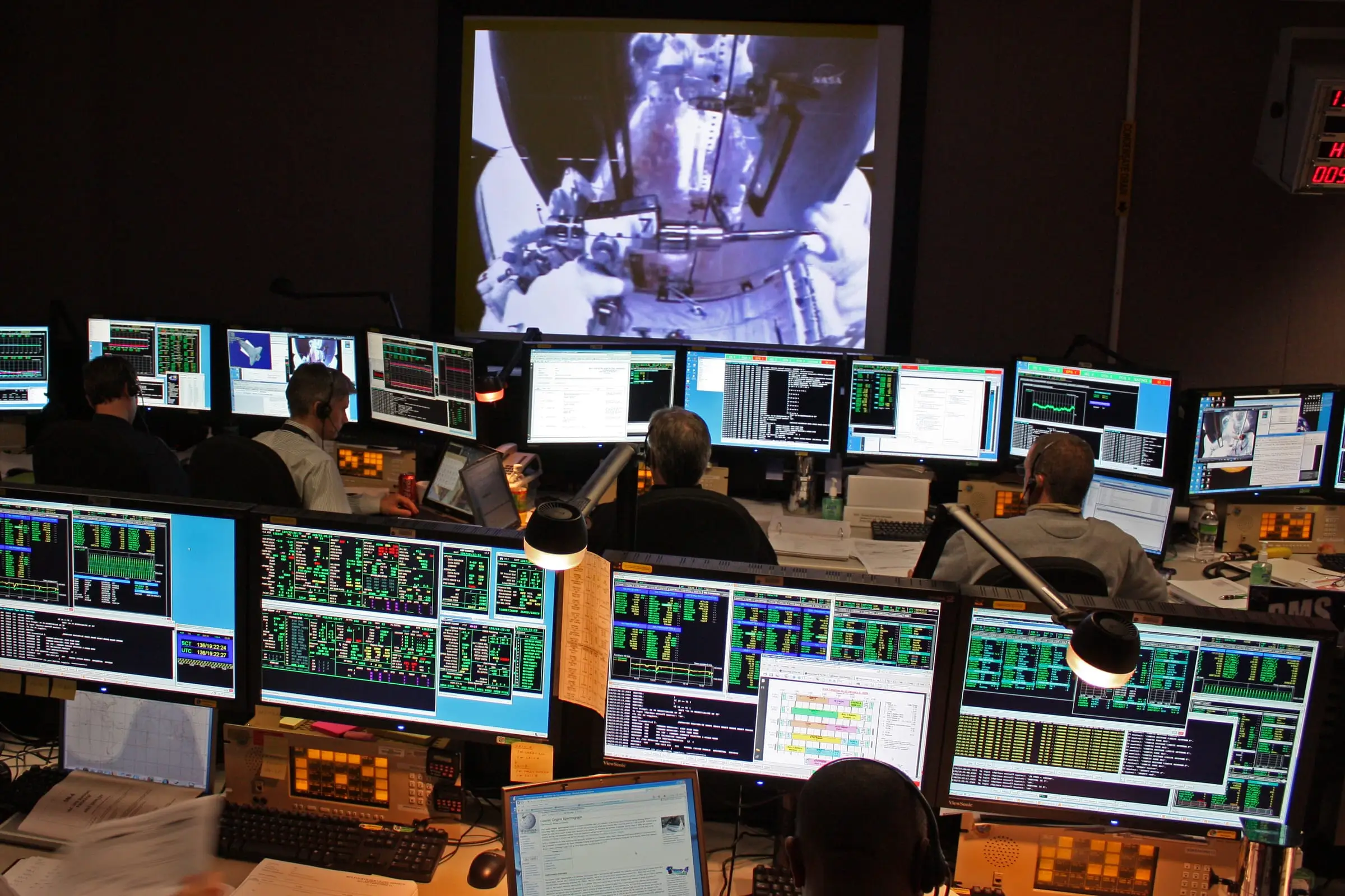 Controllers working at a row of multi-screen consoles in a dimly lit operations control center