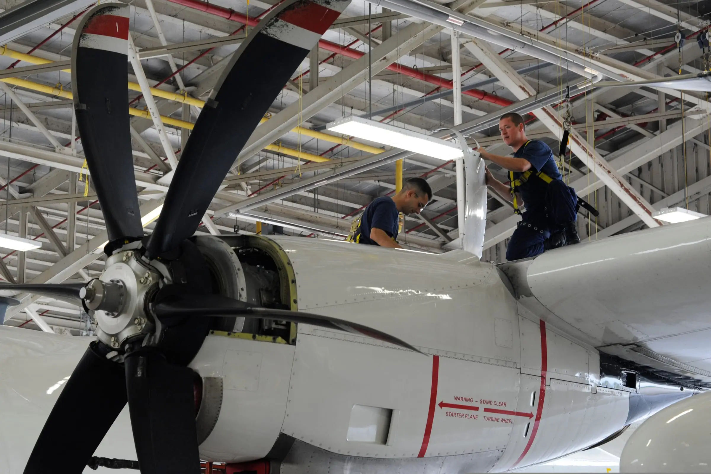 Two maintenance technicians lifting an engine cowling clear on a four-engine turboprop aircraft inside a hangar