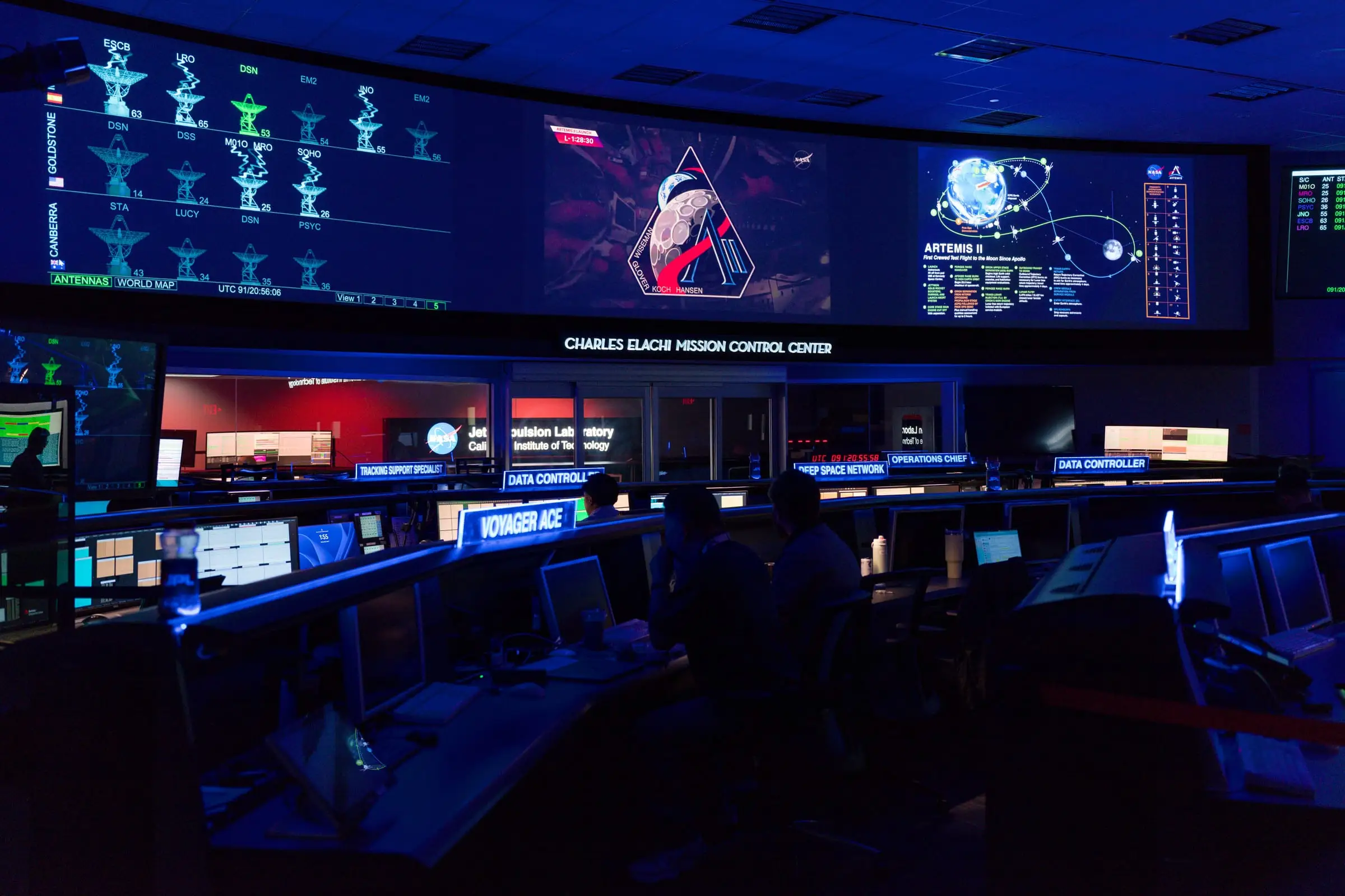 A darkened operations room lit by wall displays, with rows of consoles and staff working at them