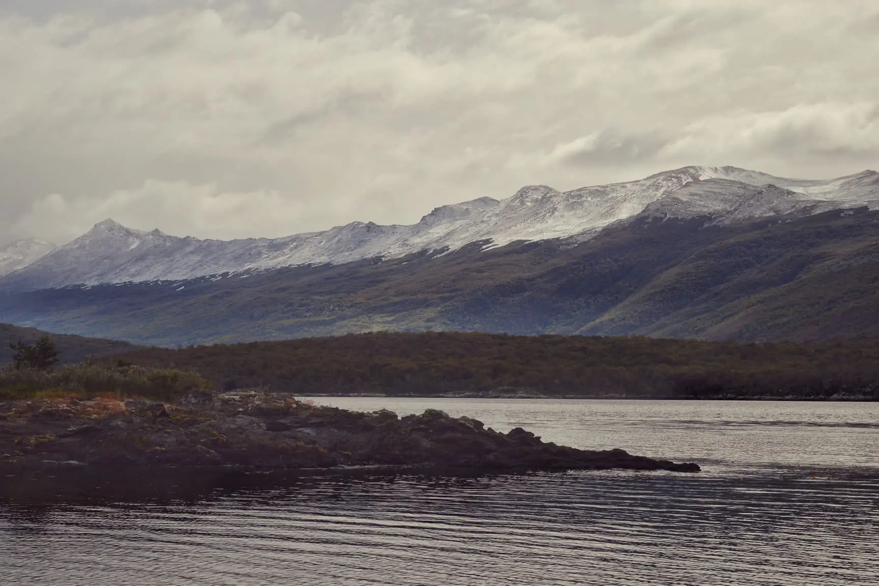 A cold-water channel between low forested headlands with a snow-streaked range behind